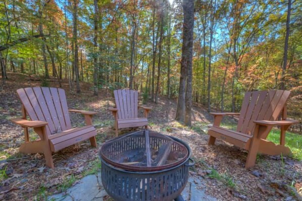 Three Adirondack chairs arranged around a fire pit in a forest setting.