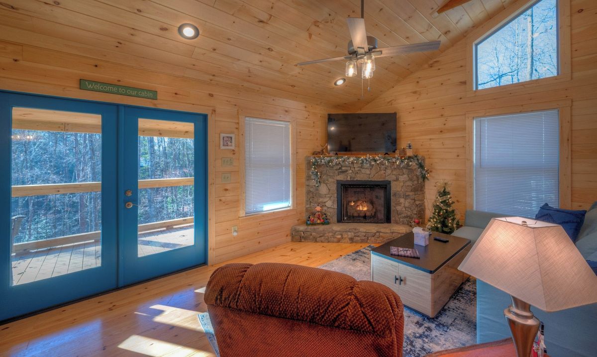 A cabin interior with a stone fireplace, a ceiling fan, and a large window overlooking a forest.
