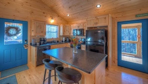 A cabin-style kitchen with wooden walls, a central island, bar stools, and a view of the forest through the windows. - Not-so-Tiny cabin. King Bed and Secure Dog Yard. (Murphy)