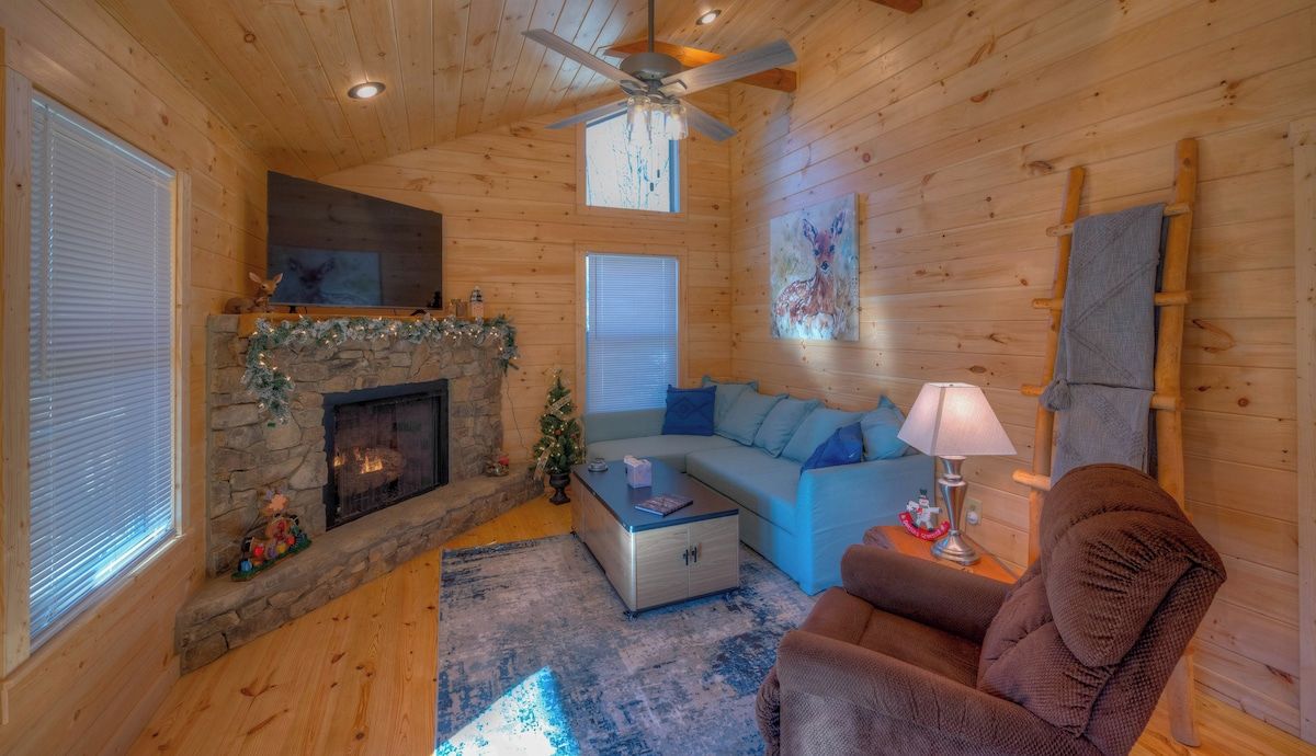 L’intérieur d’un chalet accueillant, avec une cheminée en pierre, un canapé bleu, un fauteuil marron et un ventilateur de plafond.