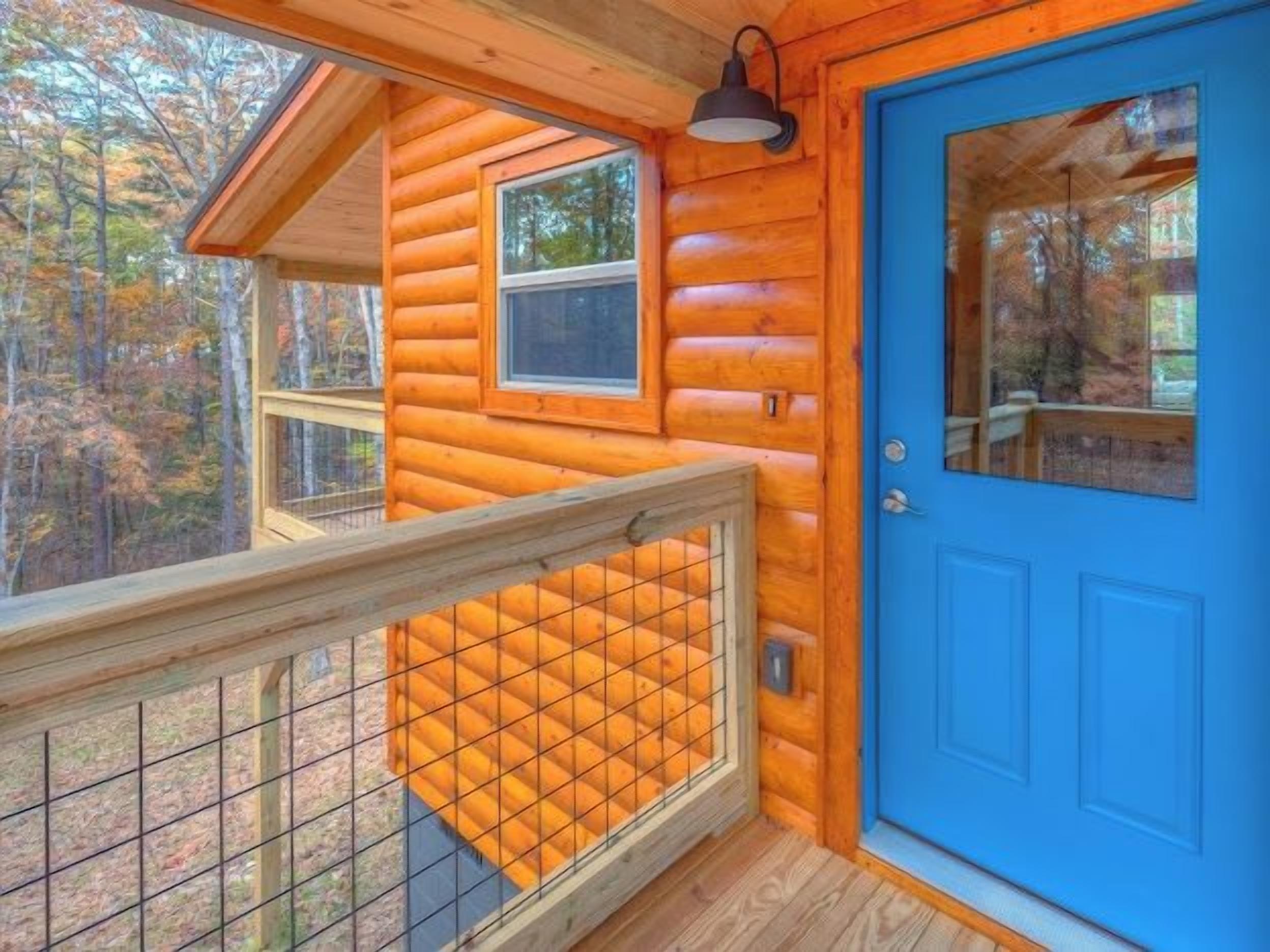 Uma cabana de tronco com porta azul, um deck de madeira com guarda-corpo e uma janela com vista para uma floresta.