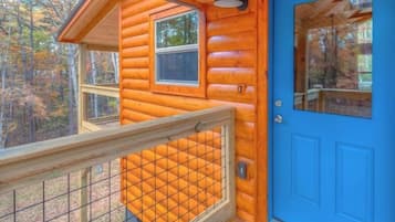 Uma cabana de tronco com porta azul, um deck de madeira com guarda-corpo e uma janela com vista para uma floresta.