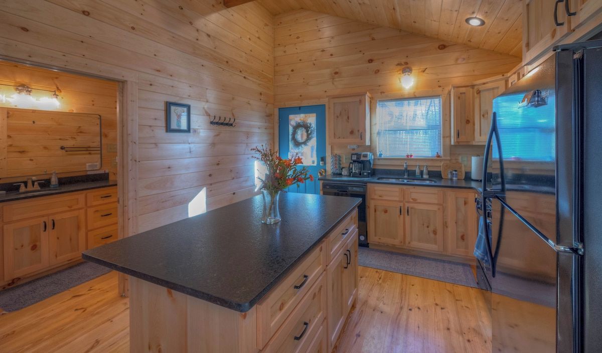 Interior de una cabaña de madera con una cocina que incluye una isla central, armarios de madera y un refrigerador.