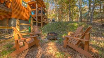 A wooden cabin with a deck, two Adirondack chairs, and a fire pit.