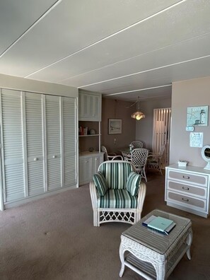 Una habitación con un sillón a rayas, un aparador blanco, una mesita de centro de mimbre y un armario con puertas batientes.