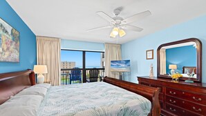 A bedroom with a bed, a dresser, a ceiling fan, a large window with curtains, and a view of the outside.