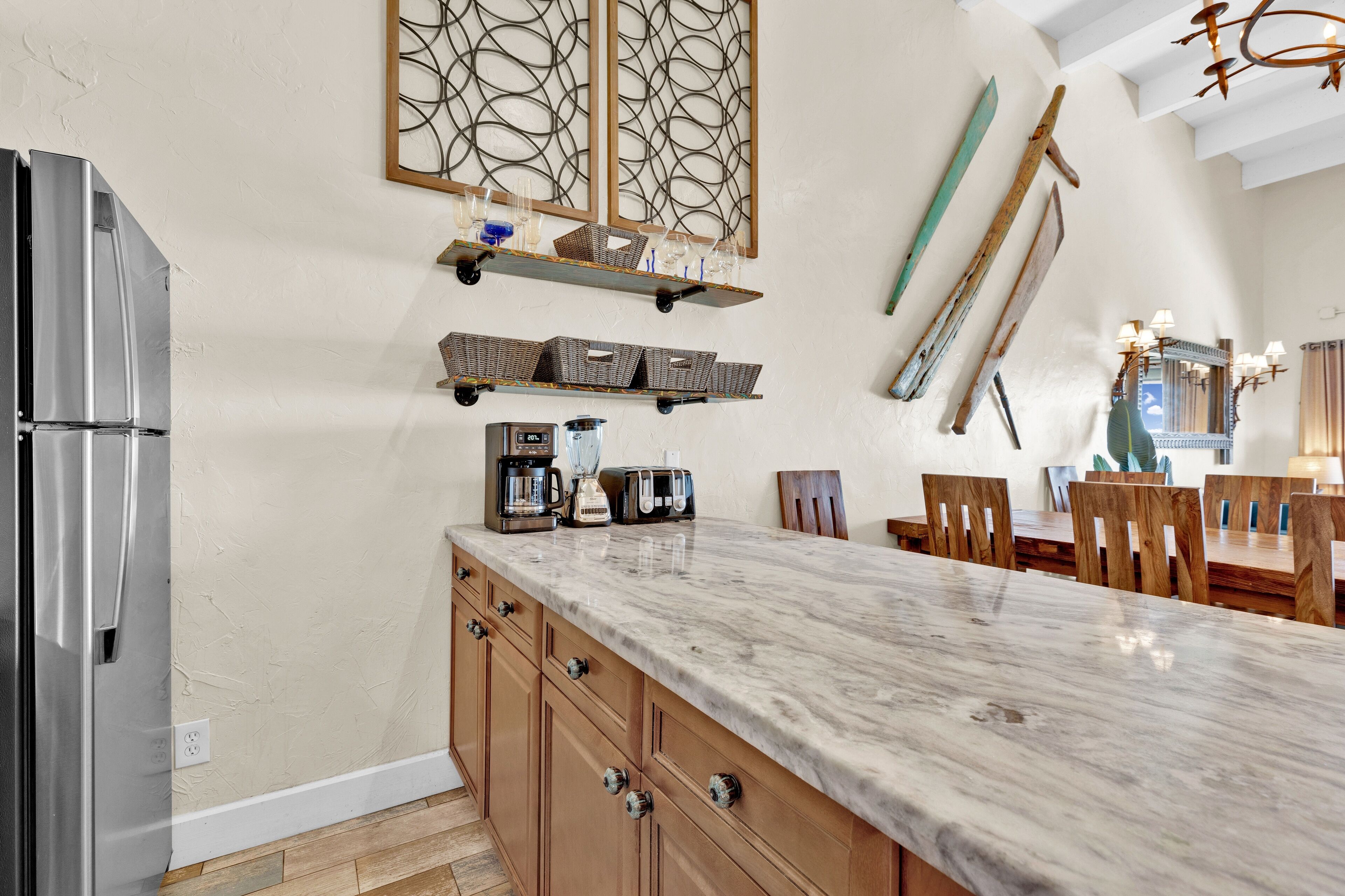 Una cocina con encimera de mármol, armarios de madera y un refrigerador empotrado. Se ven estantes con utensilios de cocina y una zona de comedor con sillas de madera.
