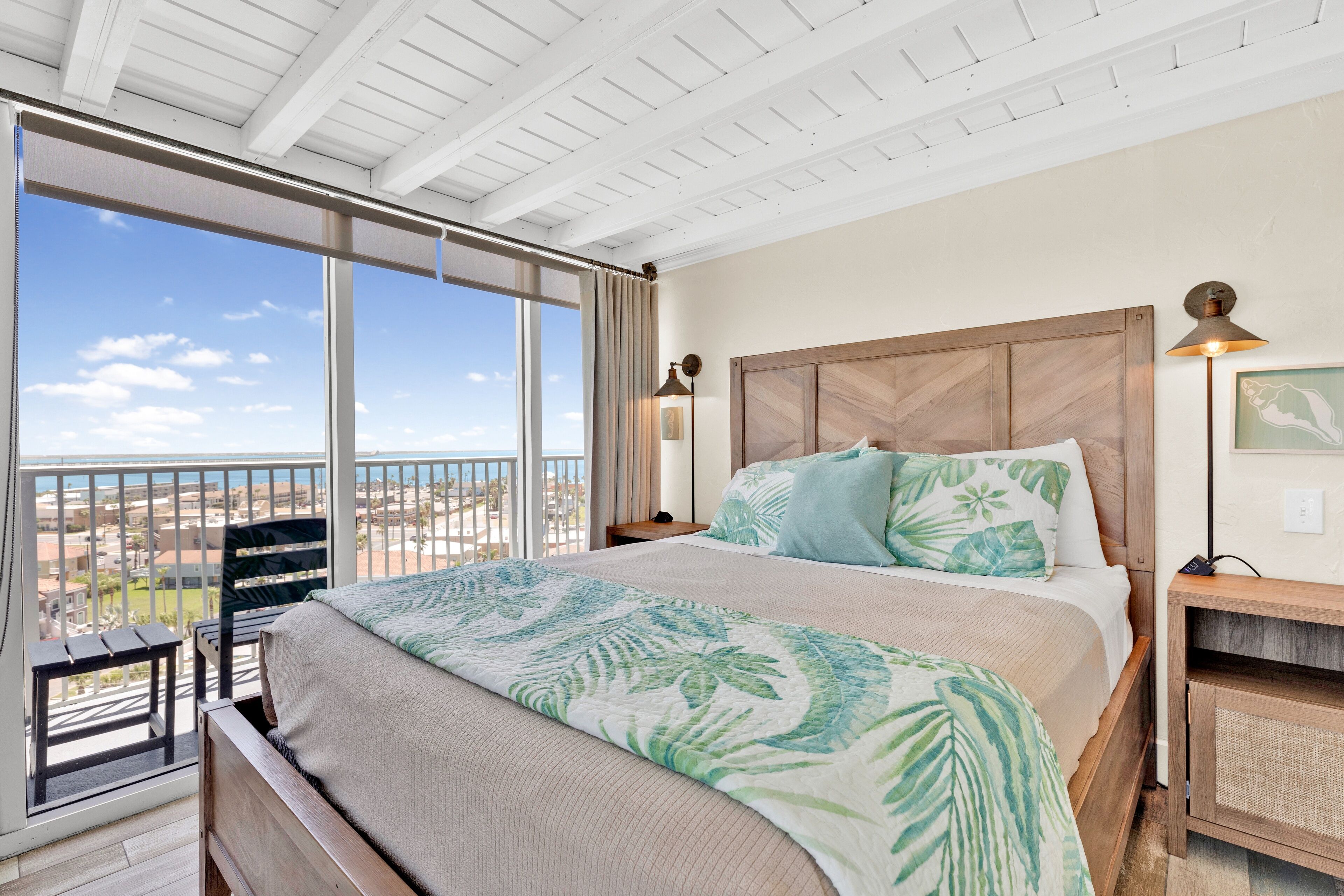 A bedroom with a large bed, a wooden headboard, a nightstand, a lamp, and a view of the city through a balcony.