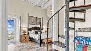 Une chambre avec un plancher en bois, un lit avec une tête de lit, une table de chevet, une lampe et une vue sur l’extérieur, visible à travers une porte.