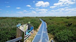 Un sentier mène à une zone de plage où se trouve une structure en bois comprenant des sièges et un mât portant un drapeau.