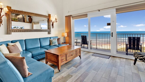 Un salon spacieux avec un canapé bleu, une table basse en bois et un grand miroir. Il est doté d’une porte-fenêtre coulissante qui donne sur un balcon offrant une vue sur l’océan.