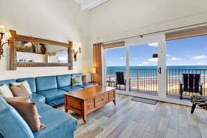 A spacious living room with a blue sofa, wooden coffee table, and a large mirror. It features a sliding glass door leading to a balcony with a view of the ocean.