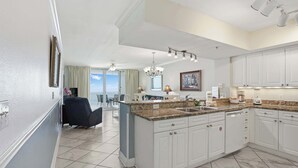 A modern kitchen with white cabinets, granite countertops, and a large island. There is a dining area with a table and chairs, and a view of the ocean through large windows.