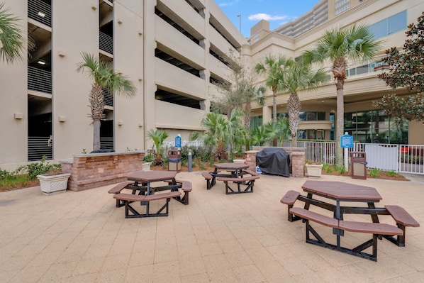 Un patio al aire libre con varias mesas de picnic, rodeado de palmeras y un edificio de varios pisos.