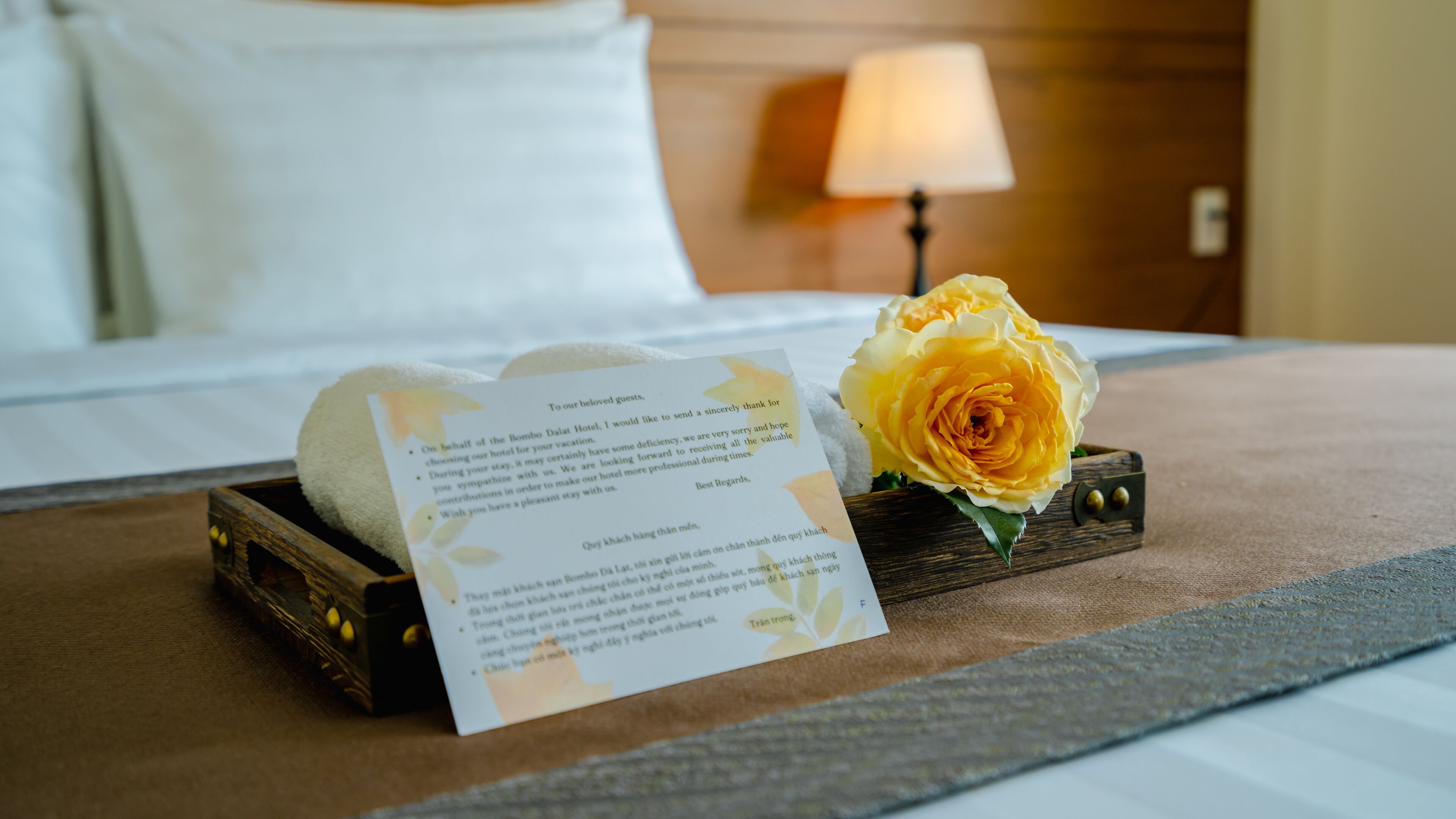 A neatly made bed with a white pillow, a wooden tray with a rolled towel and a thank you card, and a single yellow rose.
