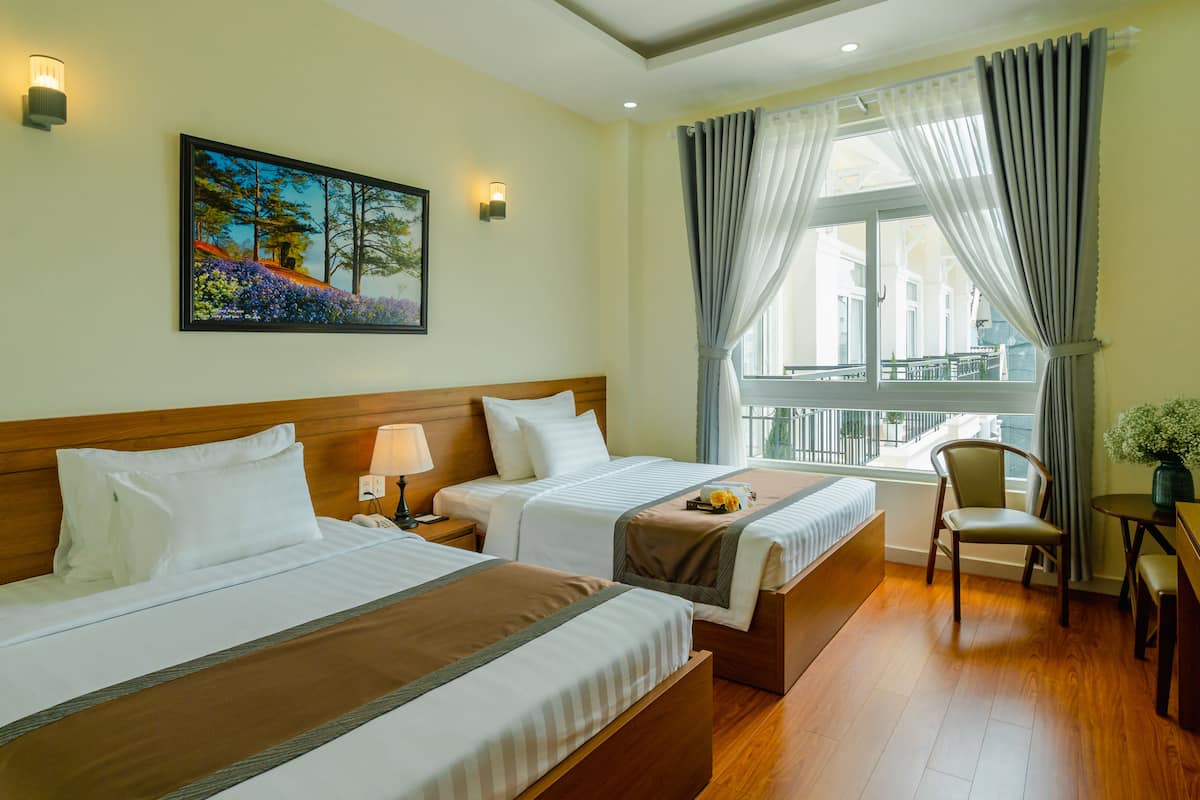 Une chambre d’hôtel avec deux lits, une tête de lit en bois, un tableau au mur, une fenêtre avec des rideaux, un bureau et une chaise.
