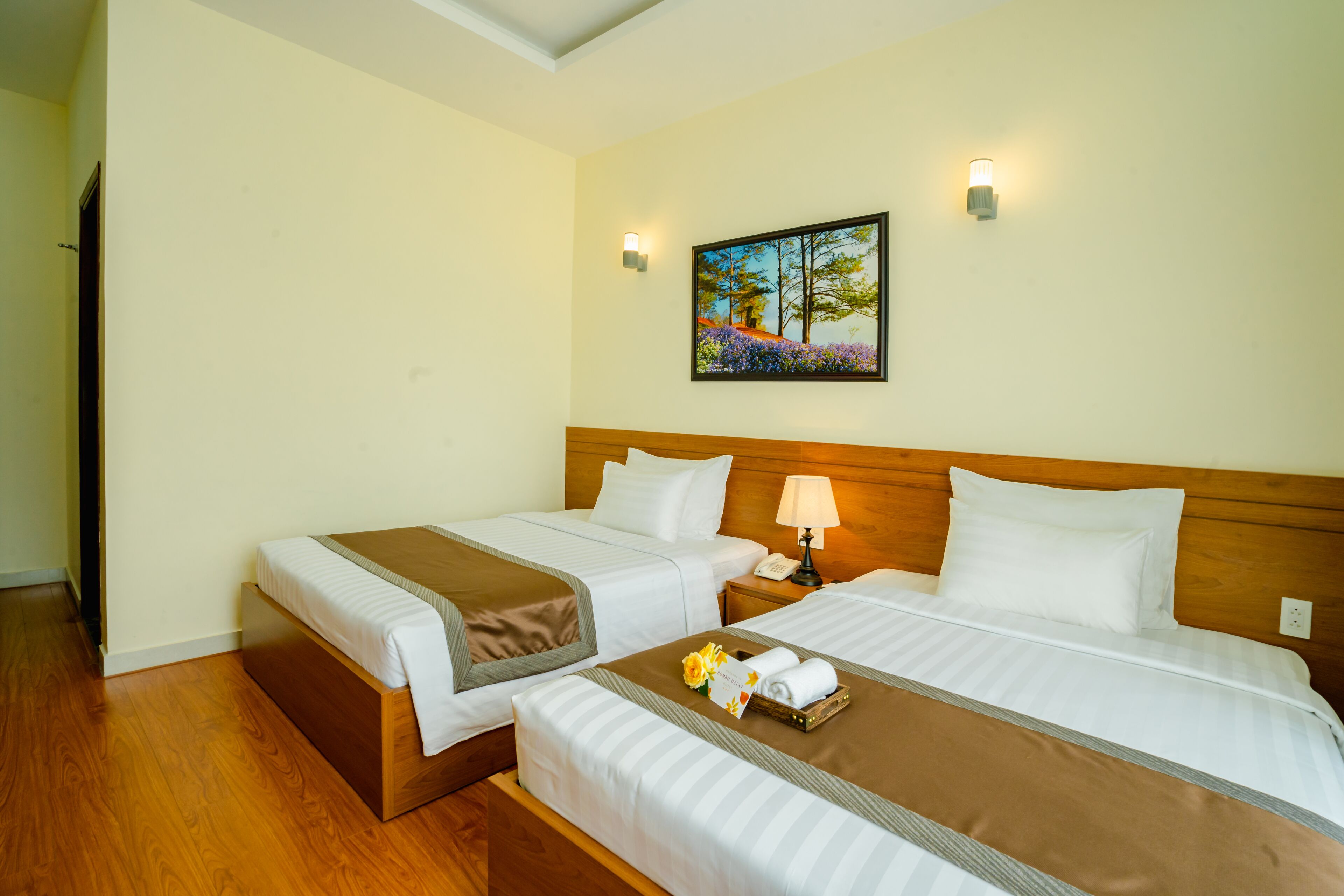 A hotel room with two beds, a wooden headboard, a nightstand with a lamp, and a painting on the wall.