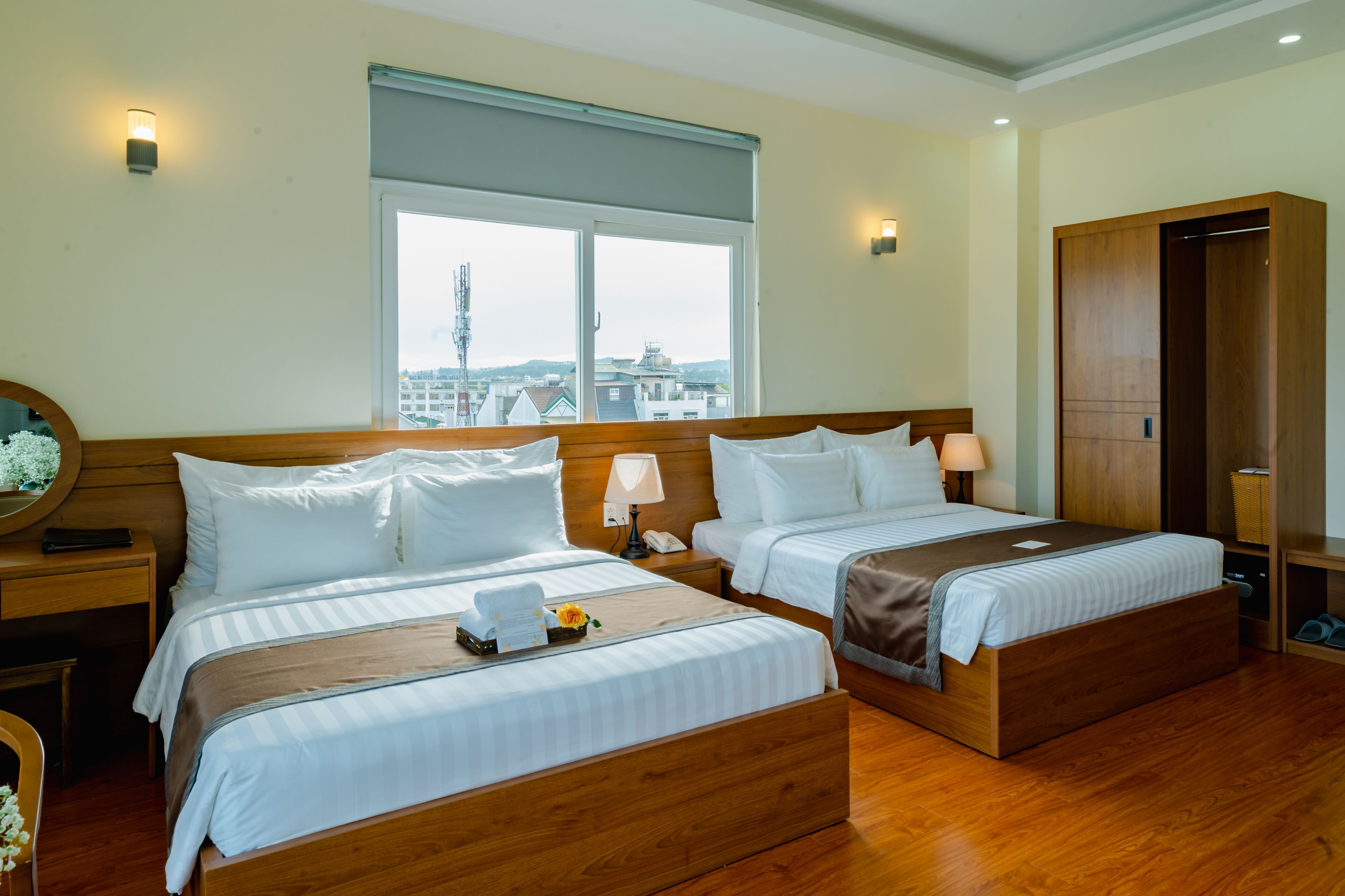 A hotel room with two beds, a large window, wooden flooring, and a wardrobe.