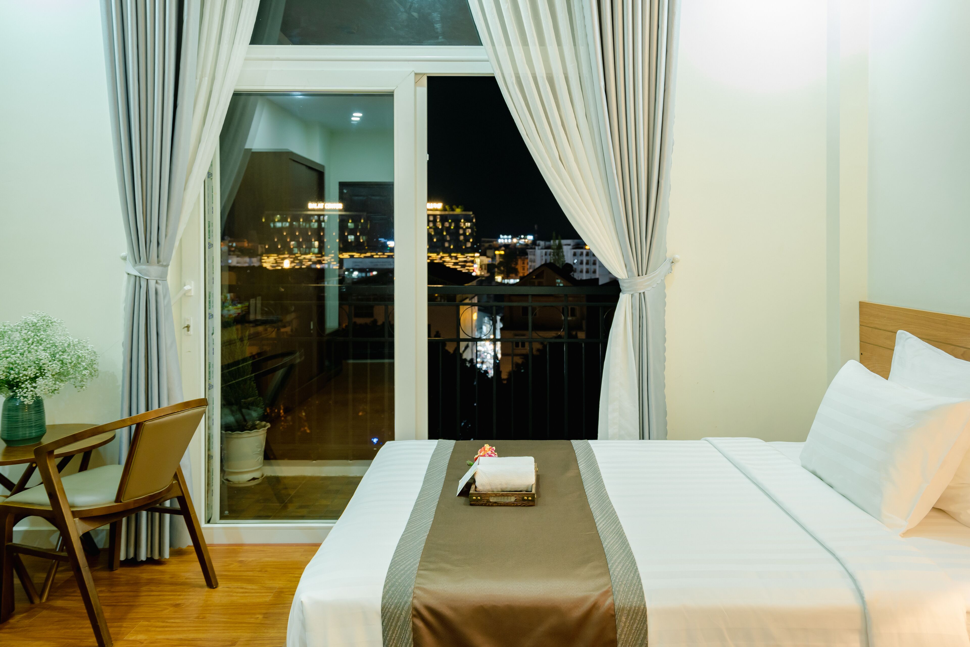 A hotel room with a bed, a chair, a table, and a view of the city at night.