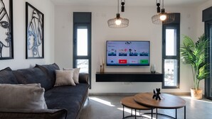 Egy modern nappali, melyben sötét színű kanapé, egy kerek, fából készült dohányasztal és egy falra szerelt, síkképernyős televízió található.