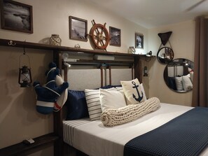 A neatly arranged bed with a striped pillow, a nautical-themed pillow, and a blanket. A plush toy is hanging on the wall, and there are framed pictures and a ship's wheel decoration above the bed. - Grace Bay beach area Apartment (Leeward Palms)