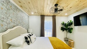 A bedroom with a wooden ceiling, a bed with white bedding and a mustard throw, a wall-mounted TV, and a large window with curtains. - Barn Loft on Gated Horse Farm (Greenville)