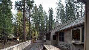 A wooden deck with picnic tables, surrounded by tall pine trees, and a building with large windows. - Big, Beautiful Golf Home, sleeps 14, situated on the golf course (Black Butte Ranch)