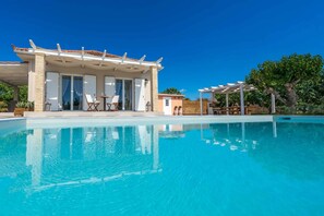 Un espace aménagé au bord de la piscine, comprenant un coin salon couvert, un petit bâtiment et une pergola.