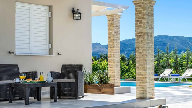 Ein überdachter Außenbereich mit Korbmöbeln, einem Tisch mit Frühstücksgebäck und Blick auf Berge sowie einen Pool.