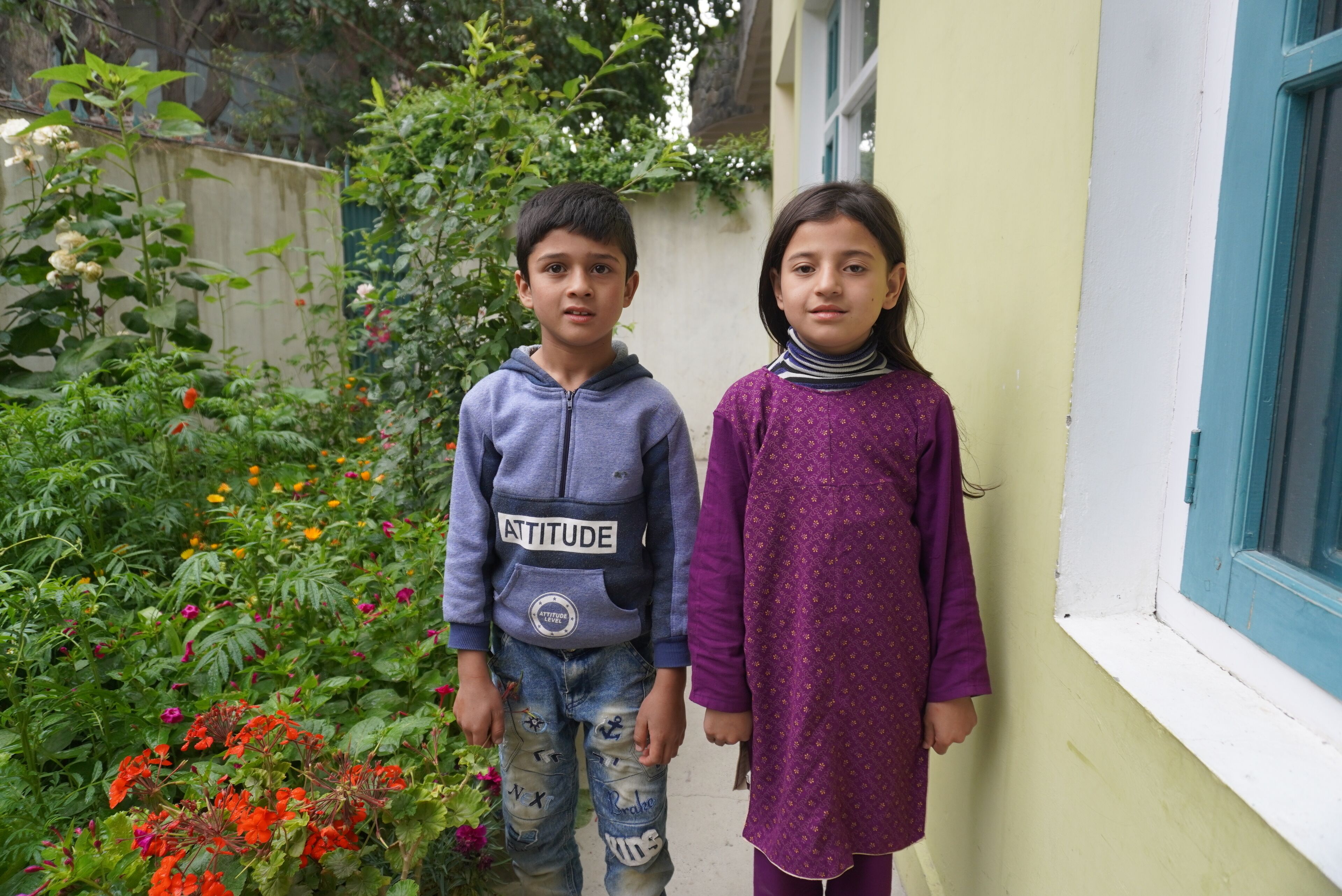 Two children standing in a garden with a variety of plants and flowers.