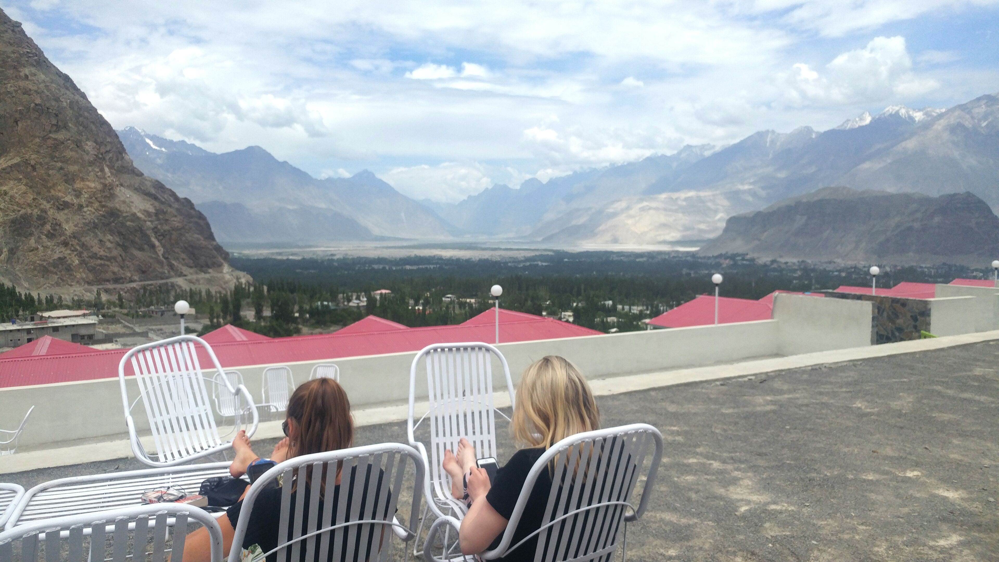 Three white metal chairs facing a mountainous landscape with a view of a valley and buildings with red roofs.