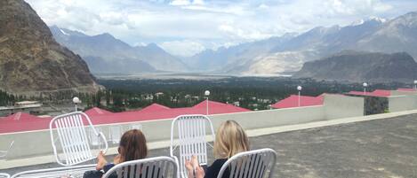 Three white metal chairs facing a mountainous landscape with a view of a valley and buildings with red roofs.