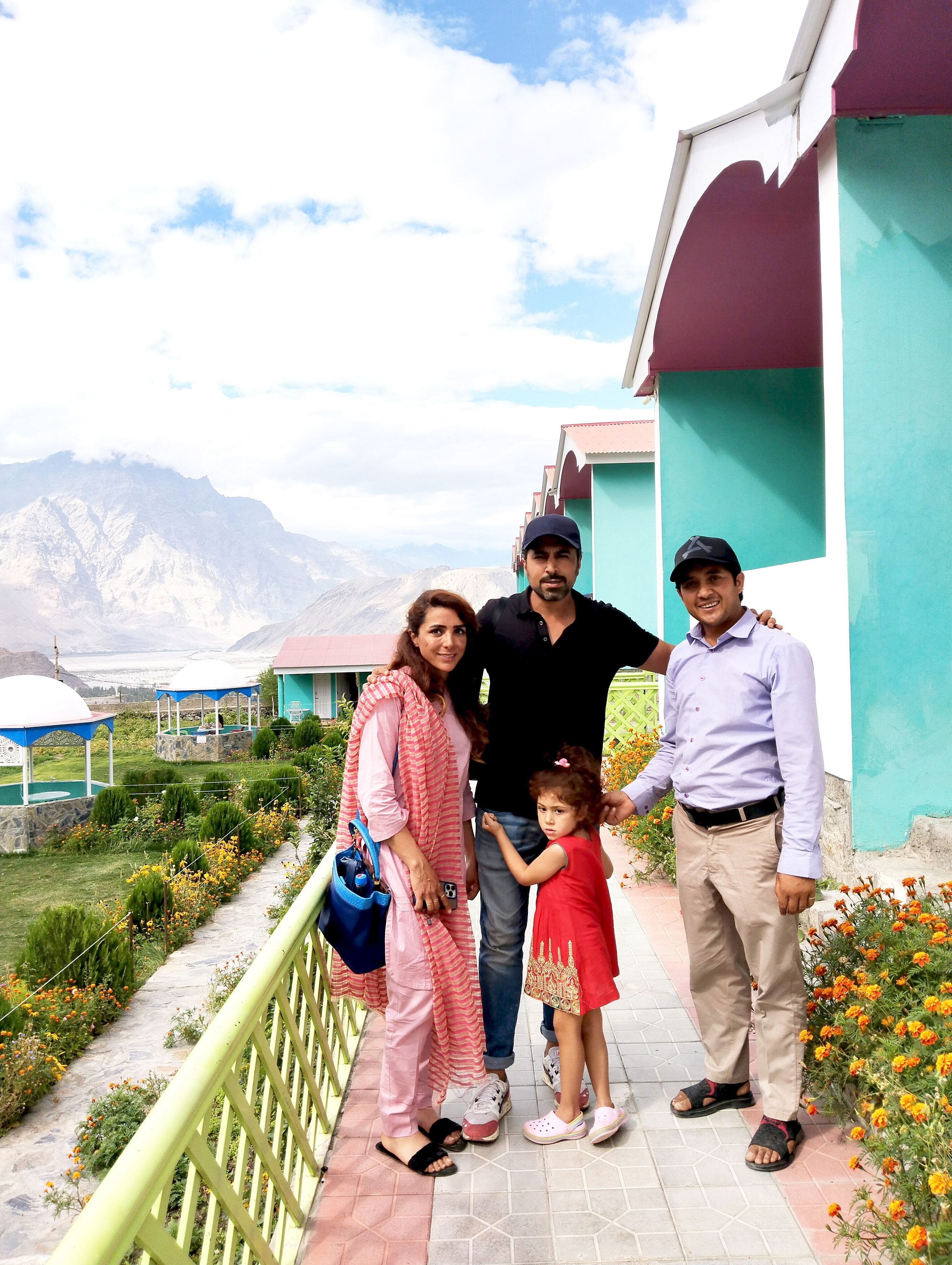 A family standing on a pathway with a mountainous backdrop, a building with teal and white walls, and a garden with flowers.