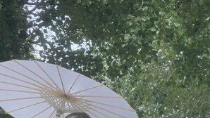 En person i en vit brudklänning som håller ett vitt parasoll och står framför en byggnad med en fontän och grönska.