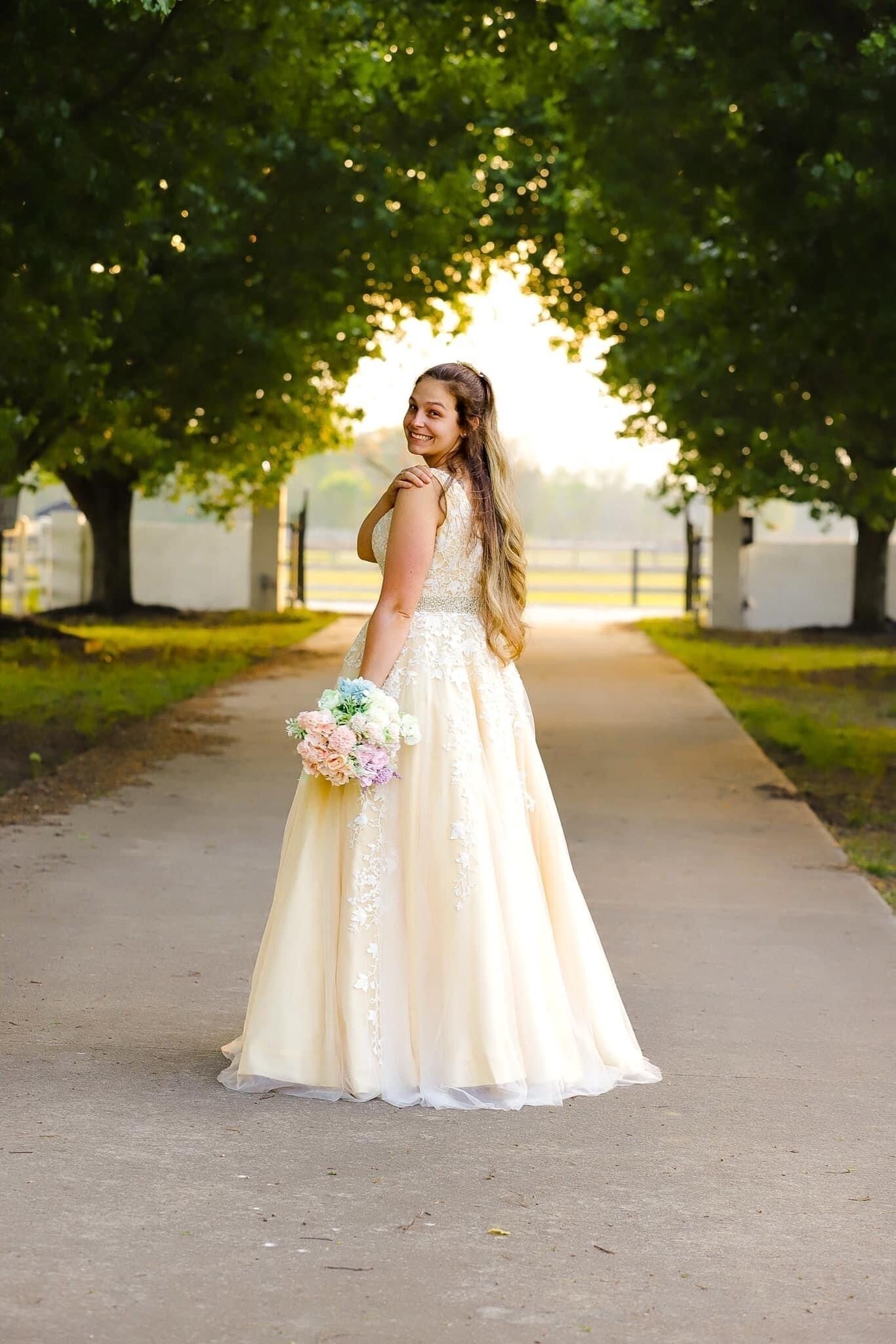 A person in a long, white dress standing on a pathway surrounded by trees.