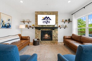 A living room with a stone fireplace, two leather sofas, a mounted TV, and a large window with a view.