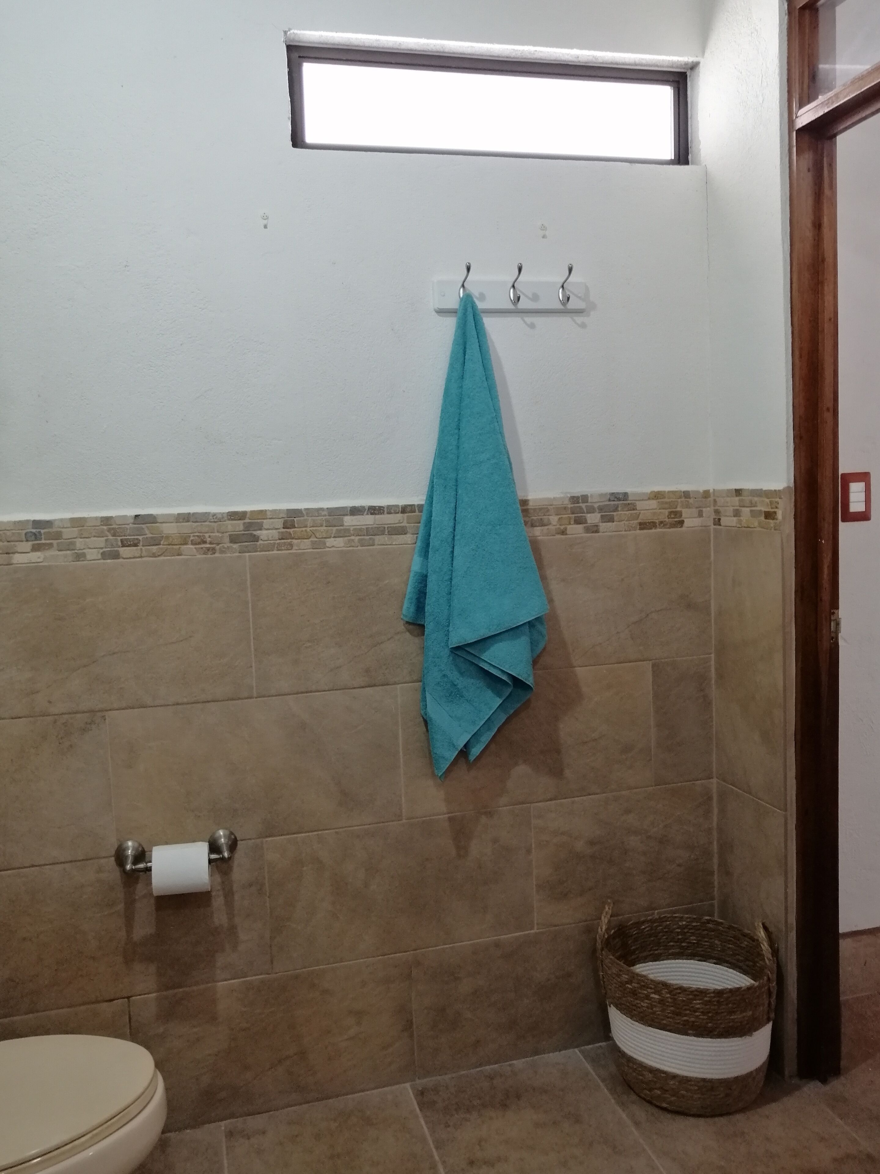 Casa de banho com sanita, um porta-toalhas com uma toalha azul, uma cesta e uma janela sobre a pia.
