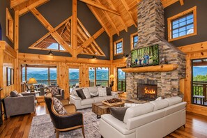 A spacious living room with a stone fireplace, wooden beams, and large windows offering mountain views. - Splashing at the Bluff: Indoor Pool, Theater, Arcade, Hot Tub, Sleeps 22 (Sevier County)