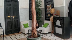A spacious lobby with a high ceiling, a large potted tree, two white sofas with green cushions, a black decorative stand, and a patterned tiled floor. - Riad étoile ocre  (Marrakech)