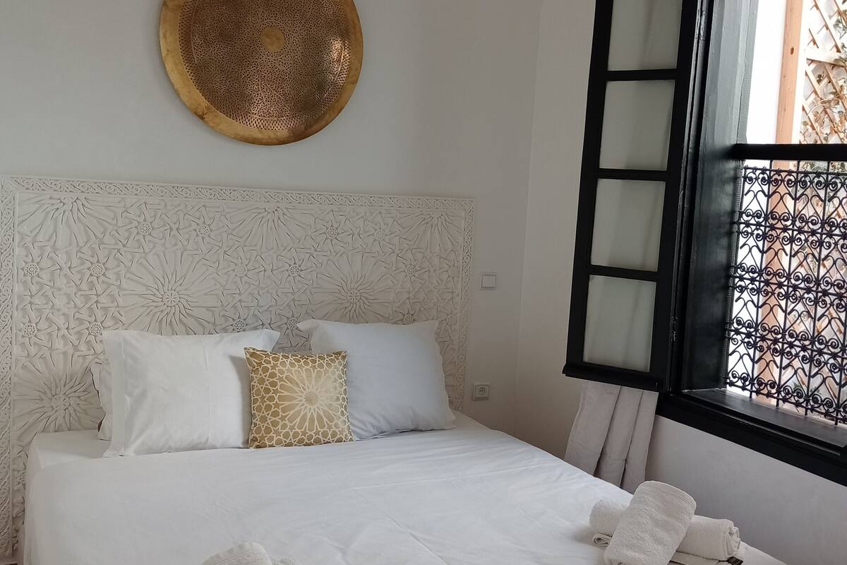 A neatly made bed with white linens and two towels, a decorative wall hanging, and a window with a black metal grille.