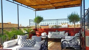 An outdoor seating area with metal tables and chairs, a covered section with a woven roof, and a view of the cityscape in the background. - Riad étoile ocre  (Marrakech)