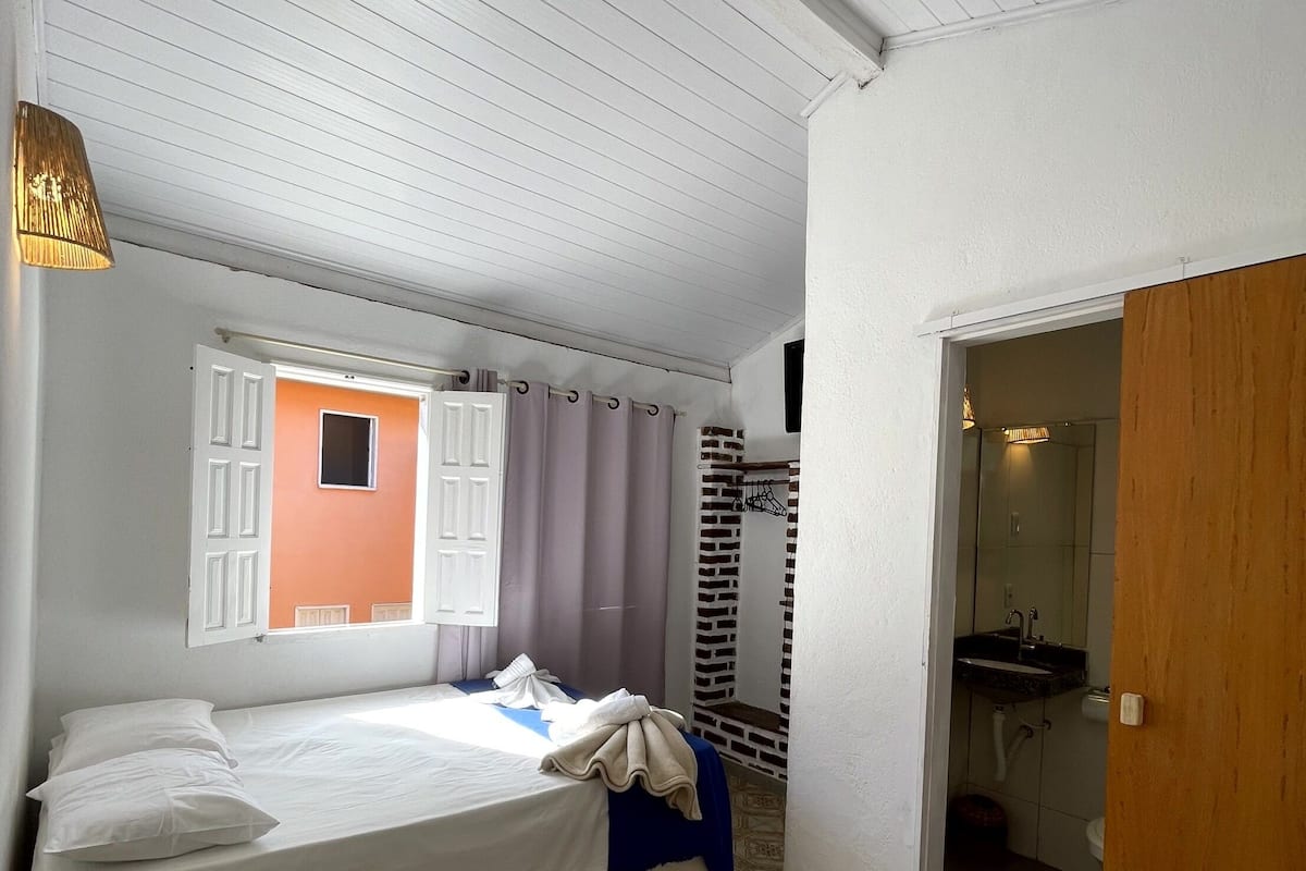 A bedroom with a bed, a desk, a lamp, a bathroom visible through an open door, and a patterned floor.