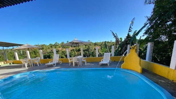 En swimmingpool med solstolar och parasoller, omgiven av en gul vägg och grönska.