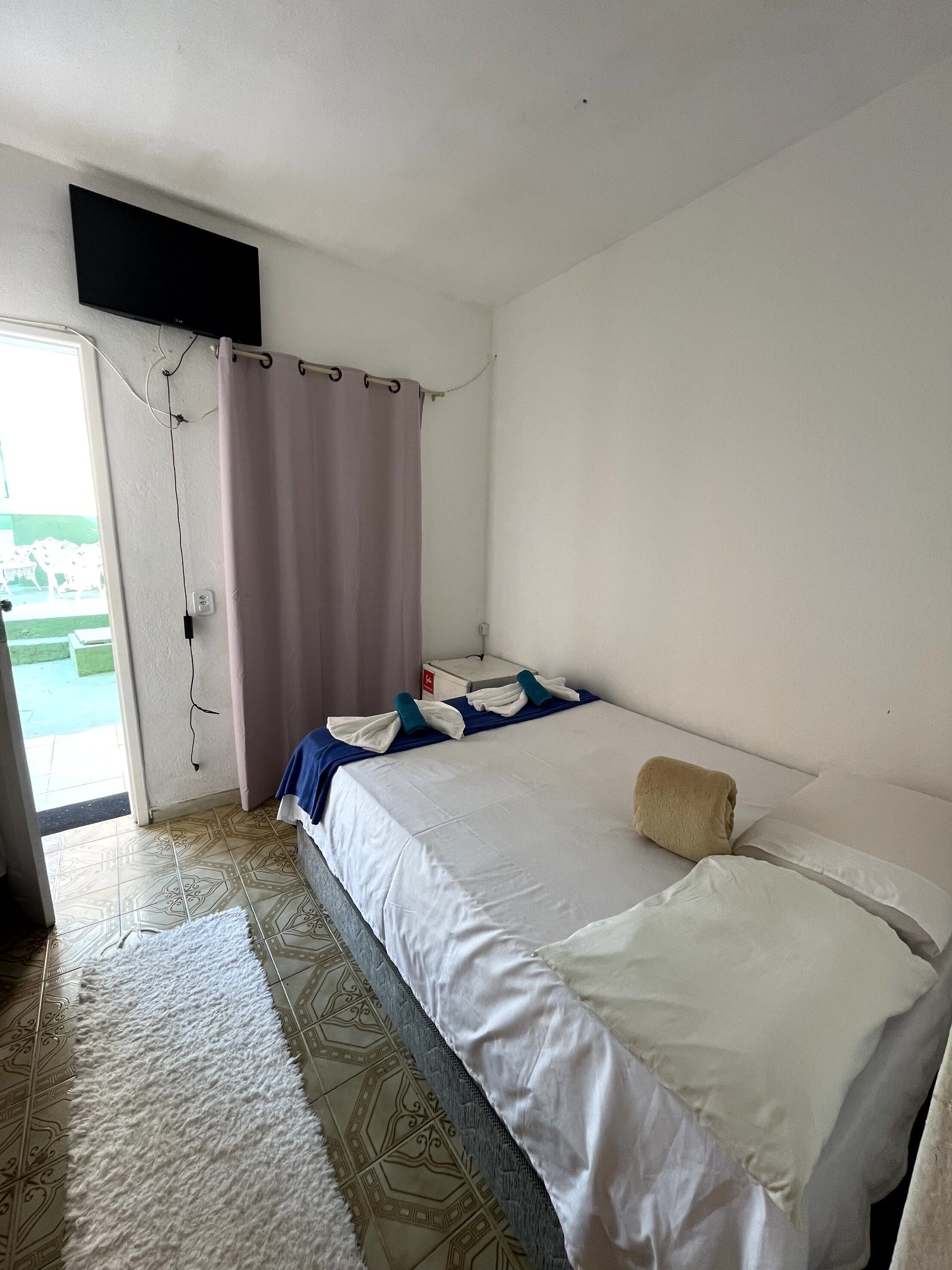 A hotel room with a bed, a TV mounted on the wall, a curtain, and a tiled floor.
