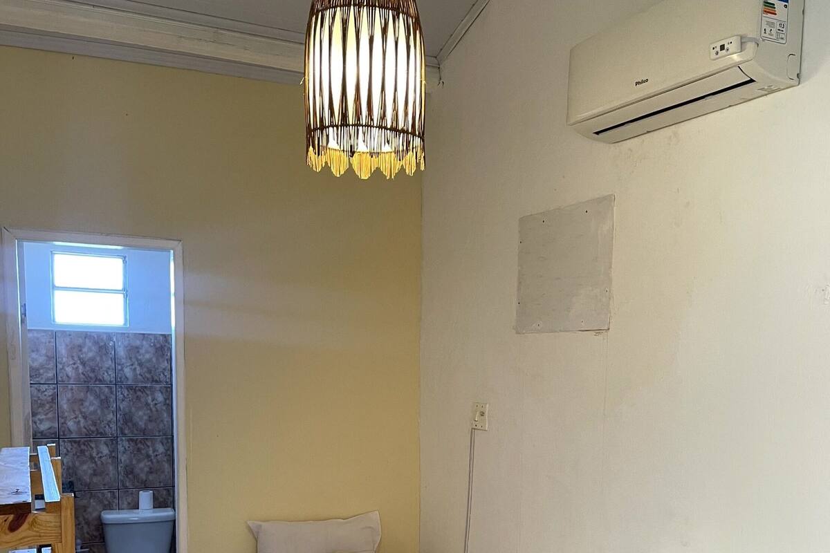 A hotel room with a bed, a wooden side table, a hanging light fixture, a bathroom with a toilet and sink, and an air conditioning unit.