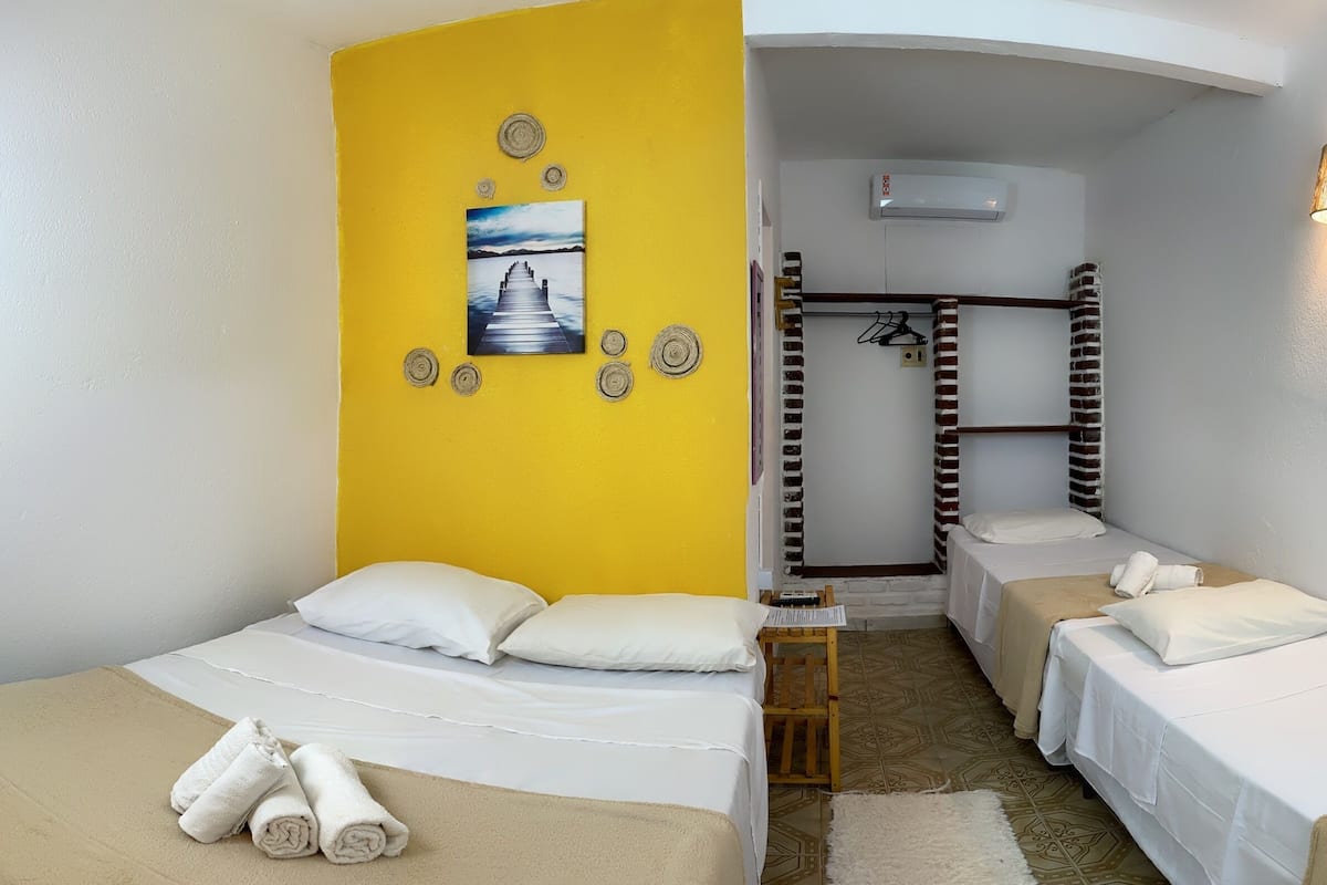 A hotel room with two beds, a yellow accent wall, a painting, a hanging light fixture, and a small table.