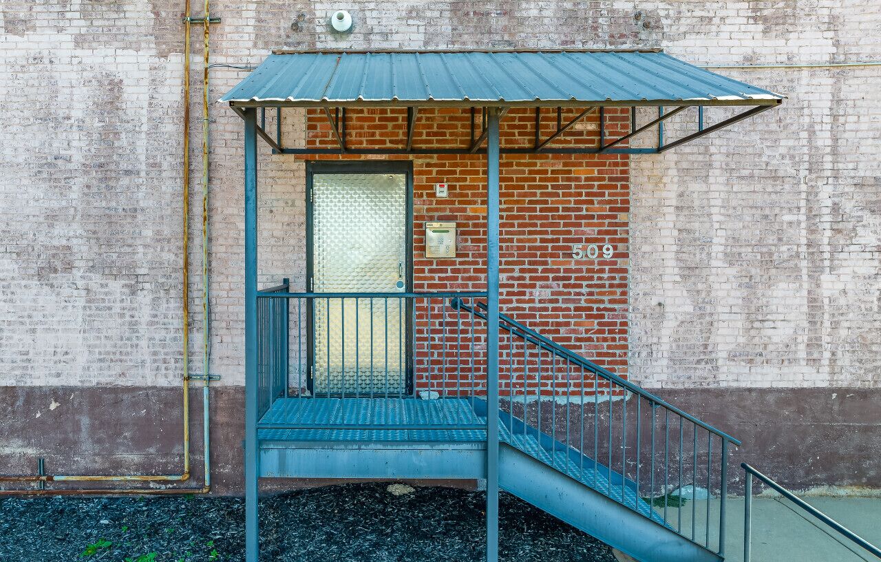 Una escalera metálica conduce a una puerta con techo metálico, el número 509 en la pared, y un muro de ladrillo al fondo.
