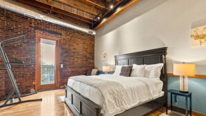A bedroom with a brick wall, wooden floor, a bed with a dark headboard, a nightstand with a lamp, and a window with a view. - Enjoy downtown living with the comforts of home! (Springfield)
