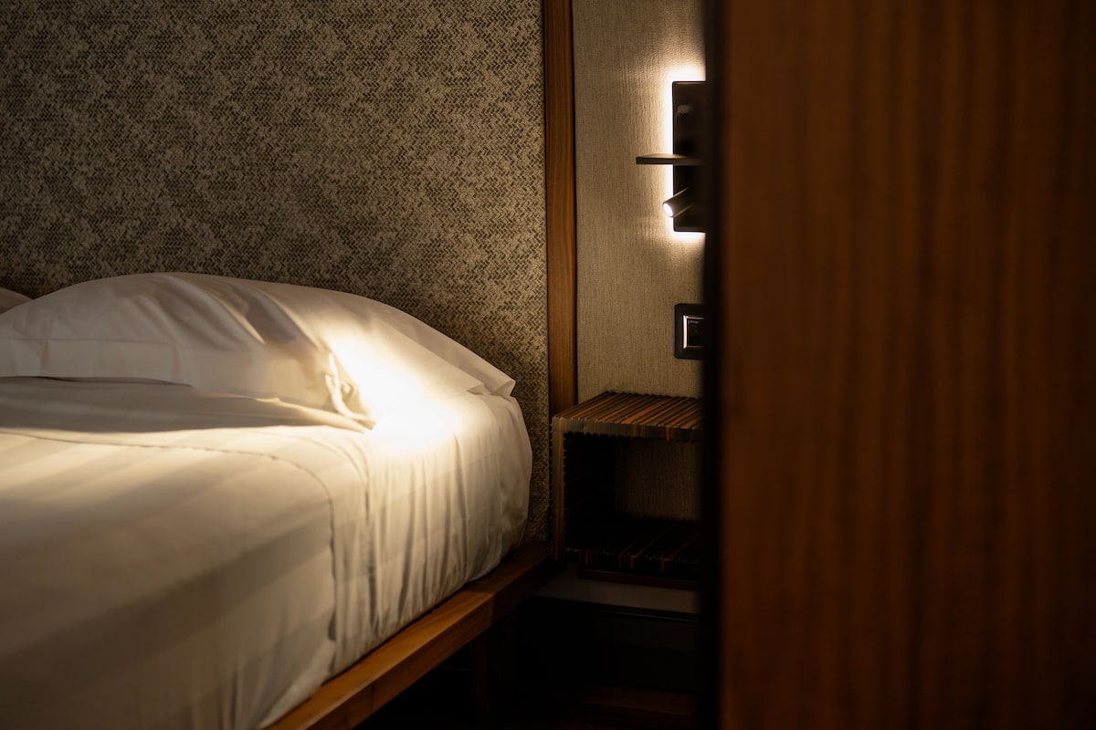 A neatly made bed with white linens in a room with a textured wall and a wooden nightstand.