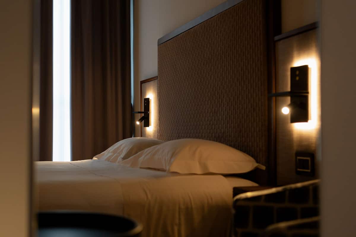 A hotel room with a bed, a nightstand, a lamp, and a view of the window with curtains.